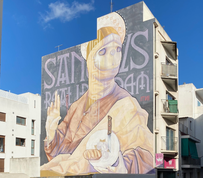 Mural of a figure resembling a saint, featuring intricate details and a knife in hand, set against a building backdrop in San Antonio, Ibiza, showcasing vibrant street art and cultural expression.