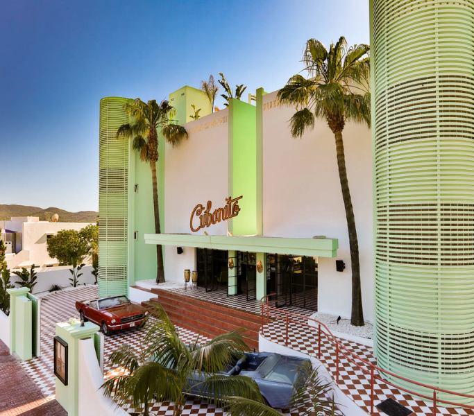 Cubanito Ibiza hotel entrance featuring vibrant green architecture, palm trees, and classic cars, showcasing luxury and Mediterranean style in a colorful setting.