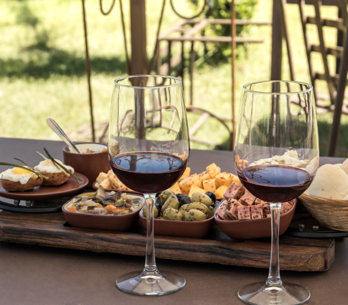 Glasses of red wine on a wooden platter with assorted cheeses, olives, and appetizers, set in a sunny outdoor dining area, highlighting Ibiza's culinary offerings.