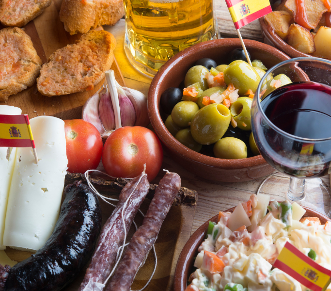 Assorted Spanish tapas including olives, tomatoes, cheese, cured meats, and a glass of red wine, with flags representing Spain, highlighting local culinary experiences in Ibiza.